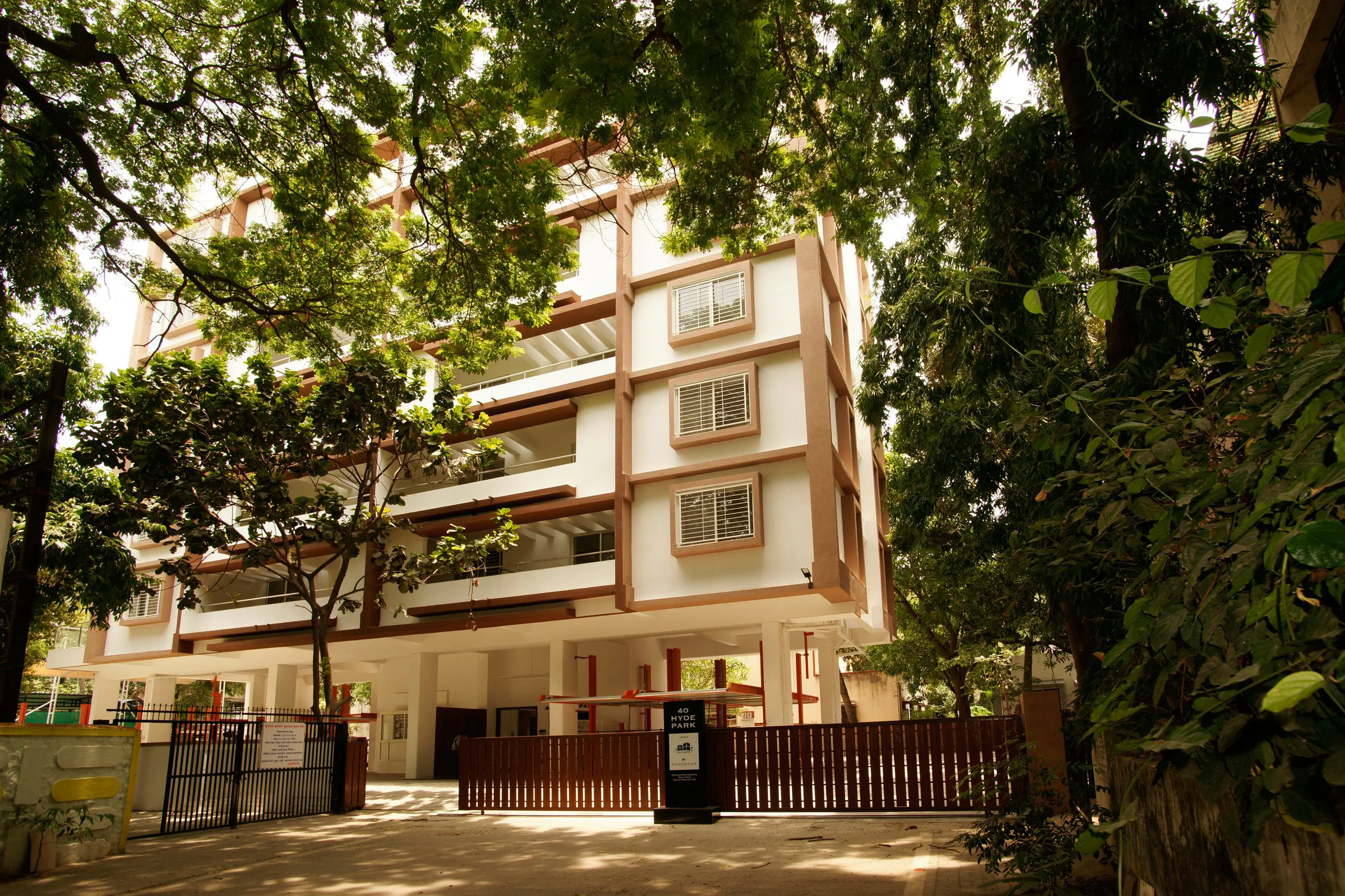 40 Hyde Park seen through mature trees: a completed six-storey building in white render with terracotta-toned framing, behind a timber gate